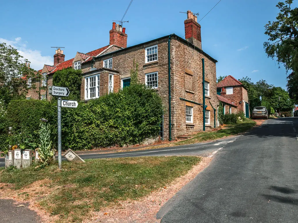 A triangle grass patch at a road junction, with a sign pointing along the road to the church. There is a house on the other side of the road on the corner.