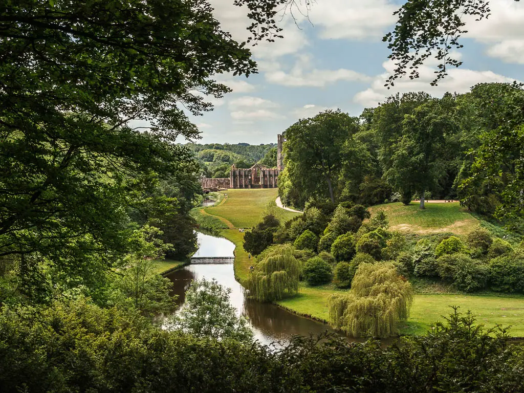 Looking down along the river, with a neatly cut green with lots of trees on the right side, and Fountains Abbey in the distance, after the walk up the hill. There are green leaved tree branches lining the left side of the frame.