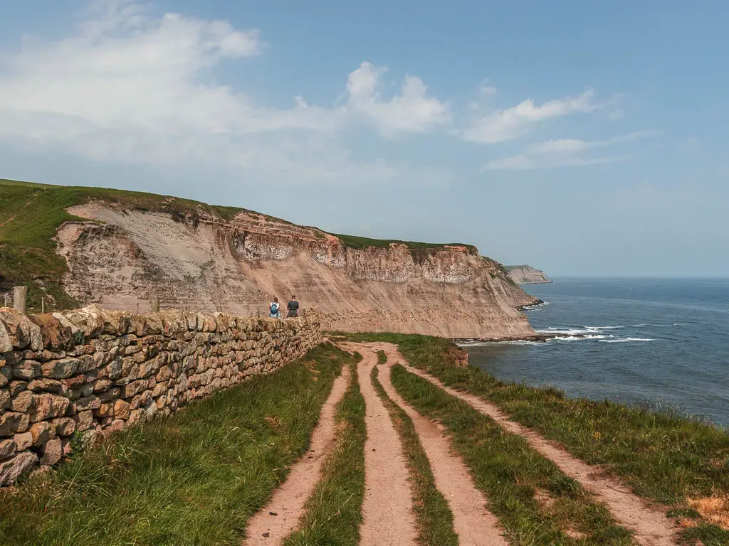 Strips of trail leading straight through the grass, with a stone wall to the left and an impressive cliffside ahead, on the coastal walk from Robin Hood's Bay to Whitby. There are two people standing past the wall ahead, looking at the cliff. The blue North Sea is to the right.