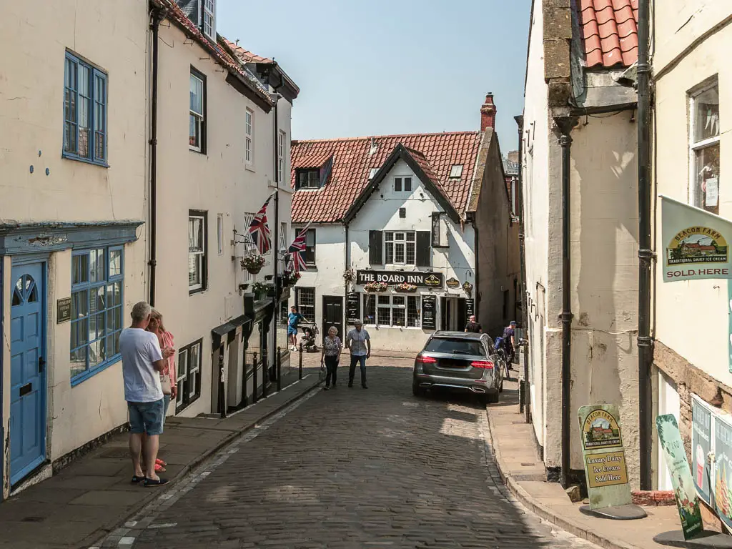 A stone road, lined with white coloured buildings, and a white walled pub at the end. There are a few people walking along the road. There is a car parked on the right.