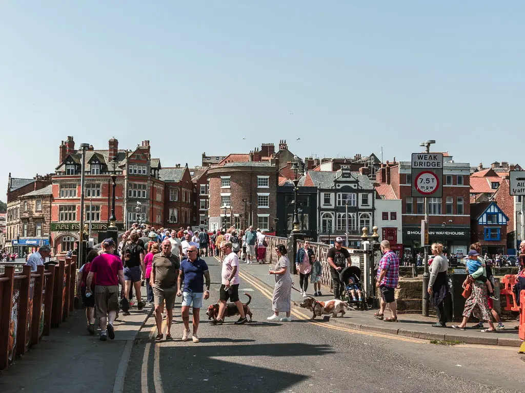 Masses of people walking along the road and over the bridge in Whitby. There are lots of buildings on the other side of the bridge.