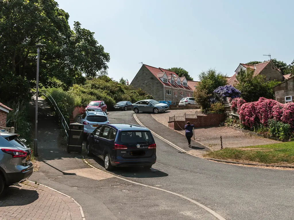 A residential street, with a footpath leading uphill to the left. There are cars parked along the street.