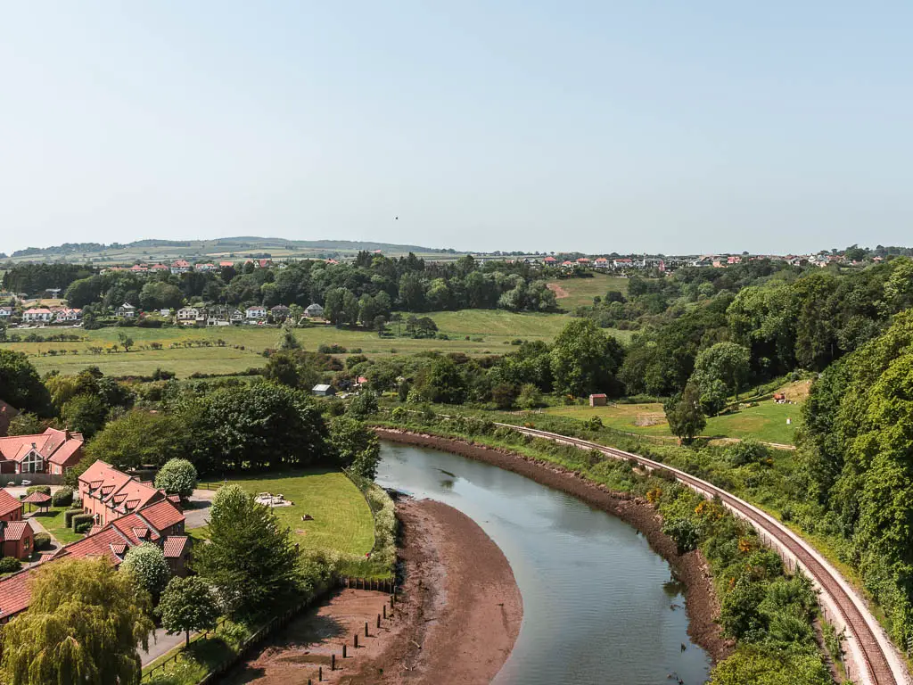 Looking down over the river Esk as it curves to the left. The railway track curves around it on the right side, and there are some red reffed houses on the left side. Across the other side of the river is fields of green with trees as far as the eye can see.