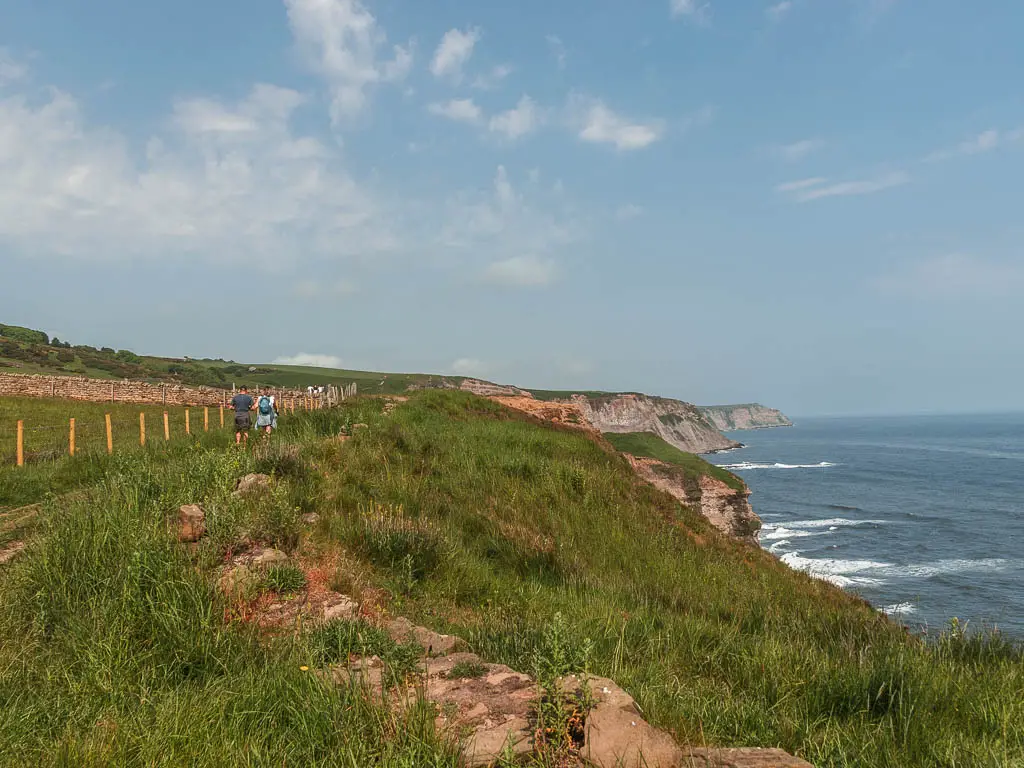 The grass clifftop, with cliff headlands below to the right, leading into the blue sea. There are a few people walking along the trail to the left.