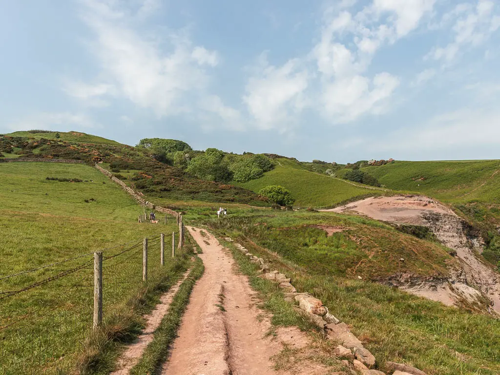 A dirt trail leading ahead, with a grass field and wire fence to the left, and undulating grass covered hilltops ahead, when walking to Whitby from Robin Hood's Bay.