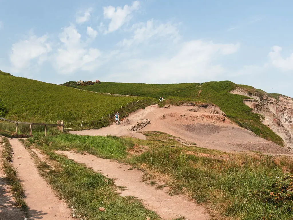 Looking over the dirt trail, to the undulating hills ahead, covered in pale dirt and grass. There are a few people walking on the hills ahead.