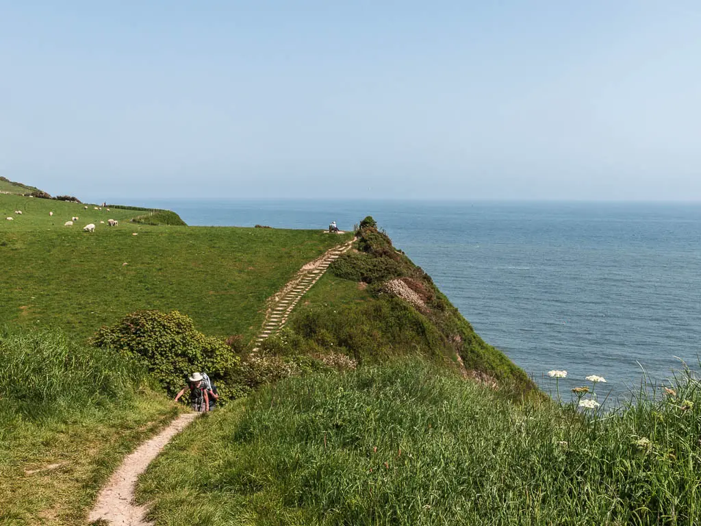 A thin path leading down the hill on the left, then up the hill in the distance. The path runs along a grass covered clifftop, with the blue North Sea to the right.