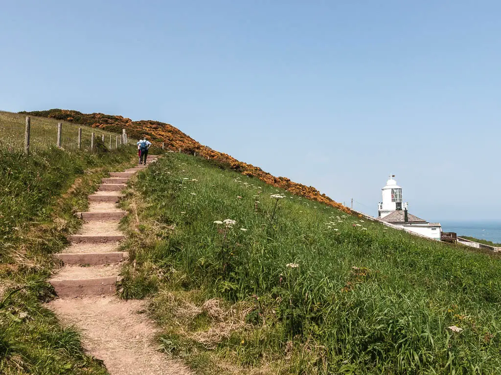 A couple of people walking up the steps on the path to the left, leading up the grassy hill, with a white lighthouse to the right, on there coastal path between Whitby and Robin Hood's Bay.