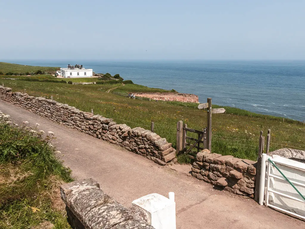 A road, lined with a stone wall. There is a grass field on the other side of the stone wall, and a white building in the distance to the left, leading towards the blue sea. There is a wooden gate in the stone wall, with a wooden footpath signpost. 