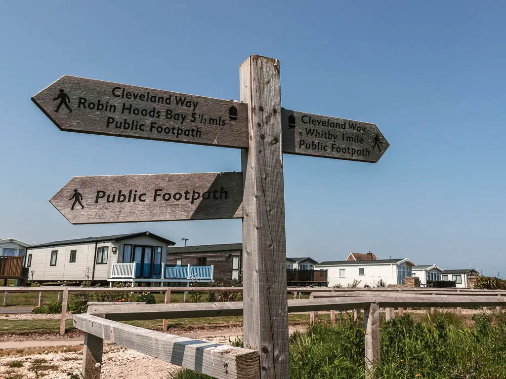 A wooden public footpath sign, pointing left to Robin Hood's Bay, and right to Whitby, partway through the walk. There are holiday caravans behind.