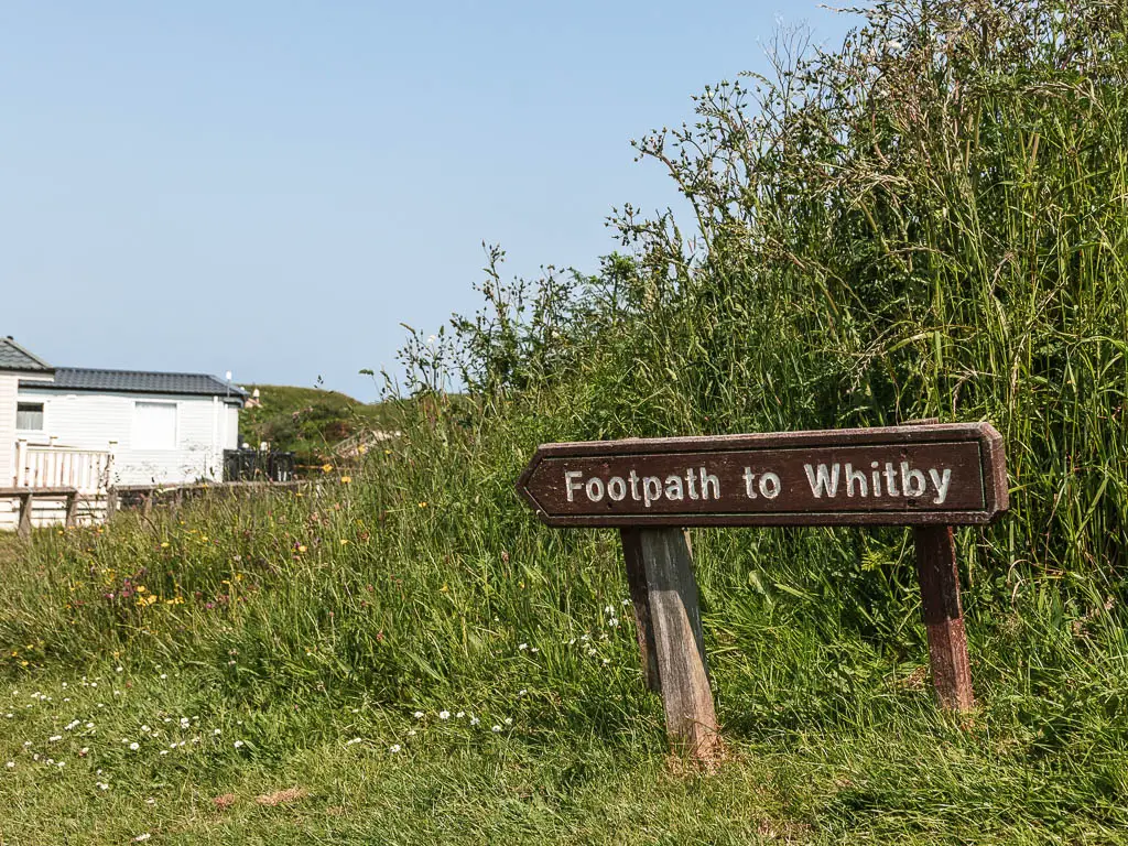 A wooden footpath sign on the grass, pointing left to Whitby.