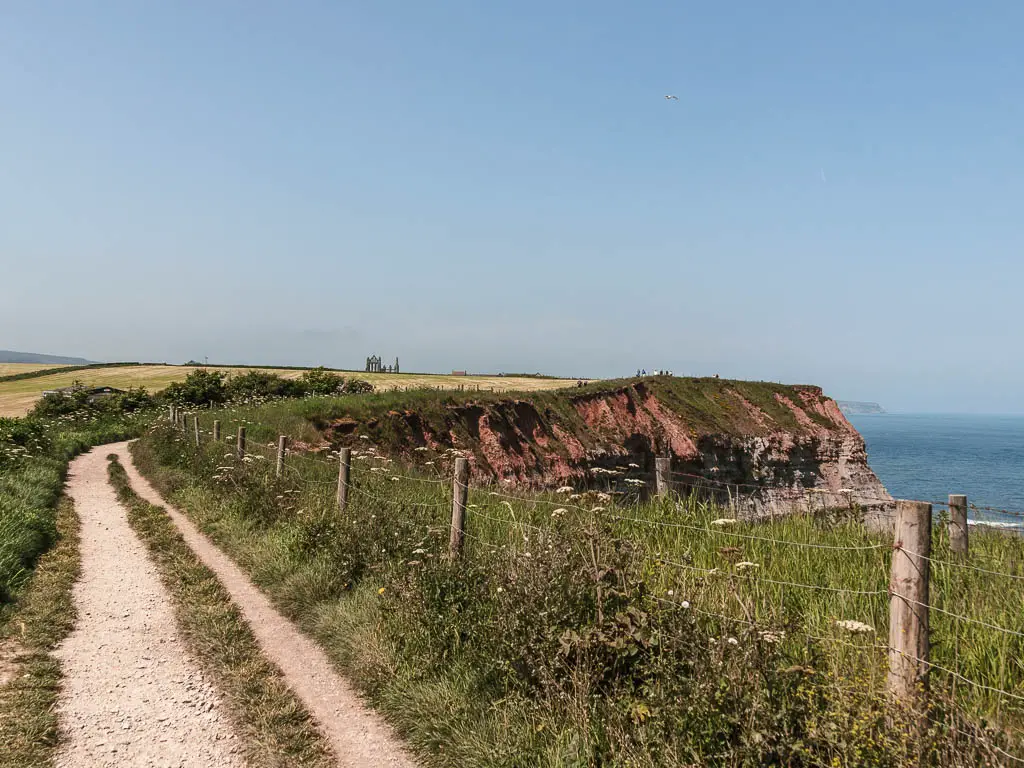 A gravel footpath on the left, with a wire fence on the right, with some tall grass on the other side, and the cliff face dropping down. Way in the distance are the ruins of Whitby Abbey.