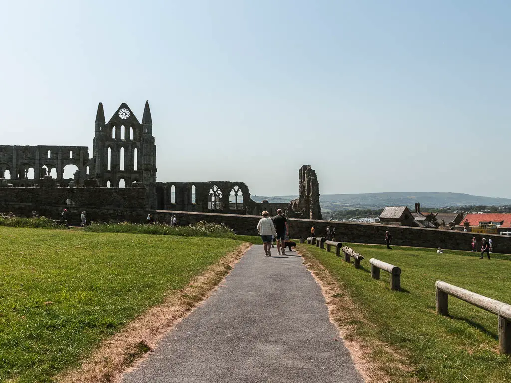 A widę footpath, leading to the ruins of Whitby Abbey straight ahead. There are a few people walking on the footpath.