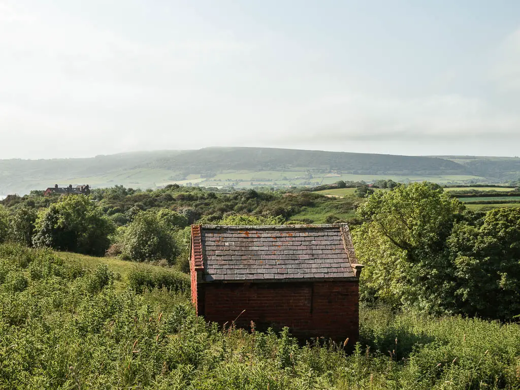 A small shed sitting on the grass hill, with a view to green grass fields with trees, as far as the eye can see, in Robin Hood's Bay, at the start of the walk to Whitby.