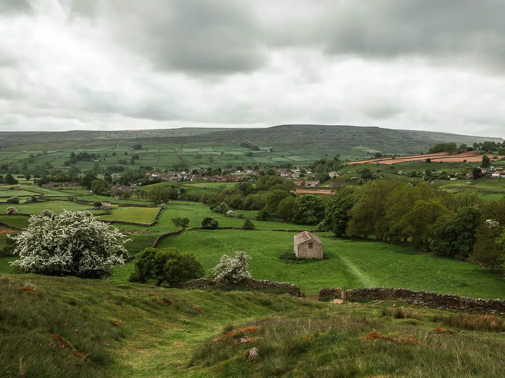 Looking down the hill at the start of the walk up to Fremington Edge. the landscape is green, with stone walls dividing the fields, and a stone shed sitting in one of the fields. The village of Reeth is visible in the distance.