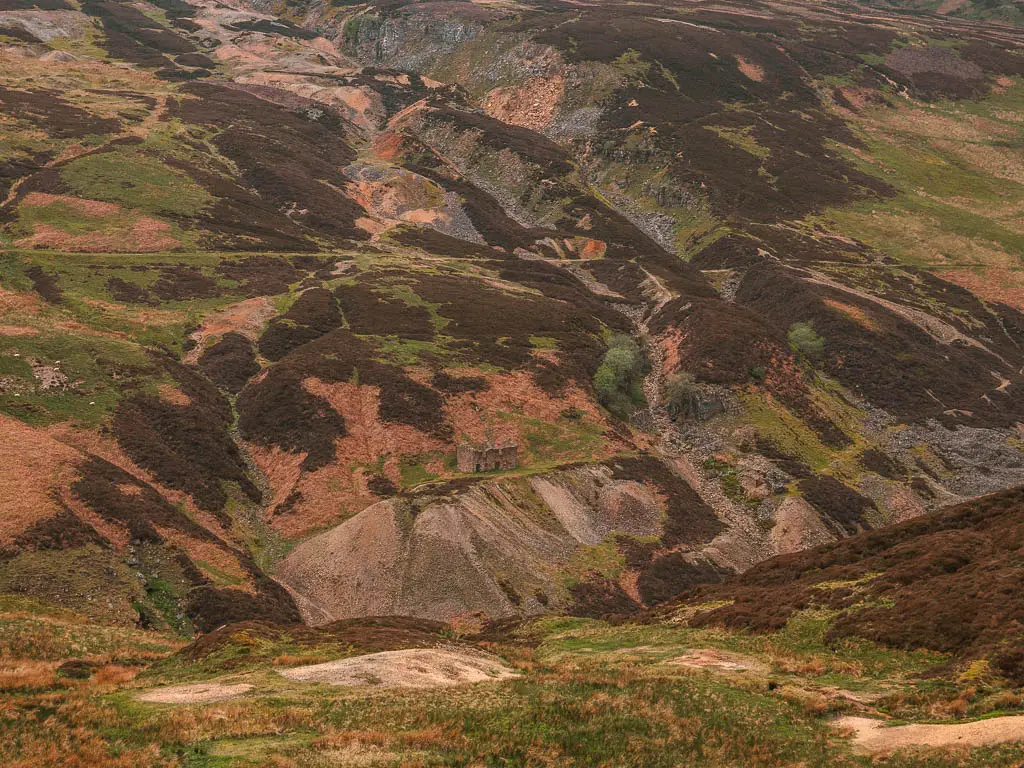 Looking down to the hillside in shades of brown, orange and green, with a stone building ruin camouflaged into the hill.