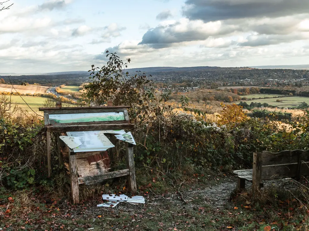 A broken information board sitting in front of a bush, looking out to a view of fields of green and trees as far as the eye can see, near the end of the walk from Merstham to Oxted. There is a wooden bench on the right, looking out across the view.