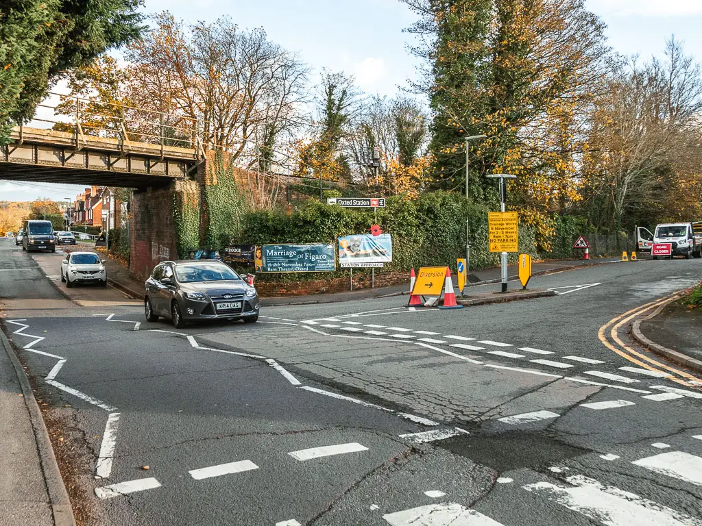 A road junction with a railway bridge ahead to the left. There is a sign by the bridge saying 'Oxted Nation'. There are a few cars driving along the road. 