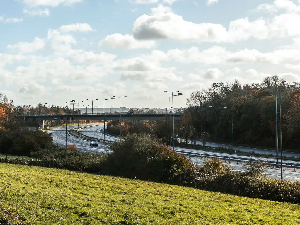 Looking across the green grass field to the M4 motorway. There are light posts lining the motorway, and a bridge across it ahead. 