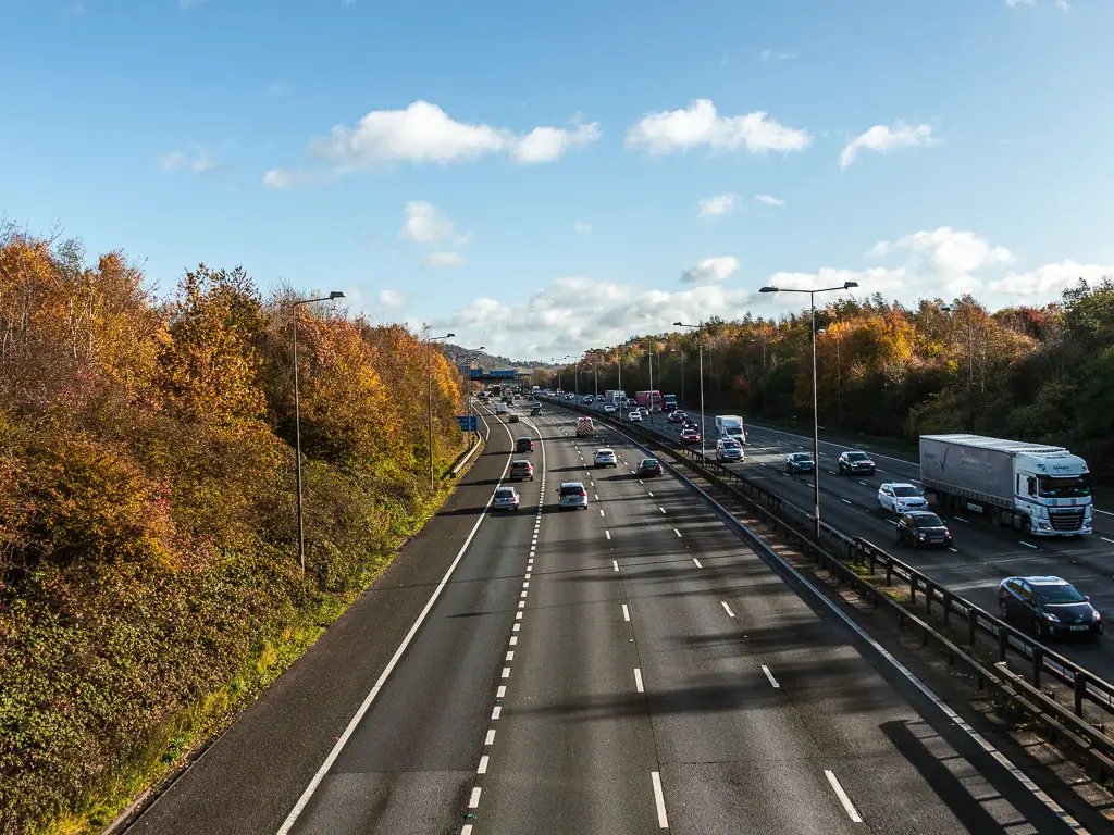 Looking down along the M4 motorway, with lots of cars driving along it. The motorway is lined with trees with orange and yellow leaves.