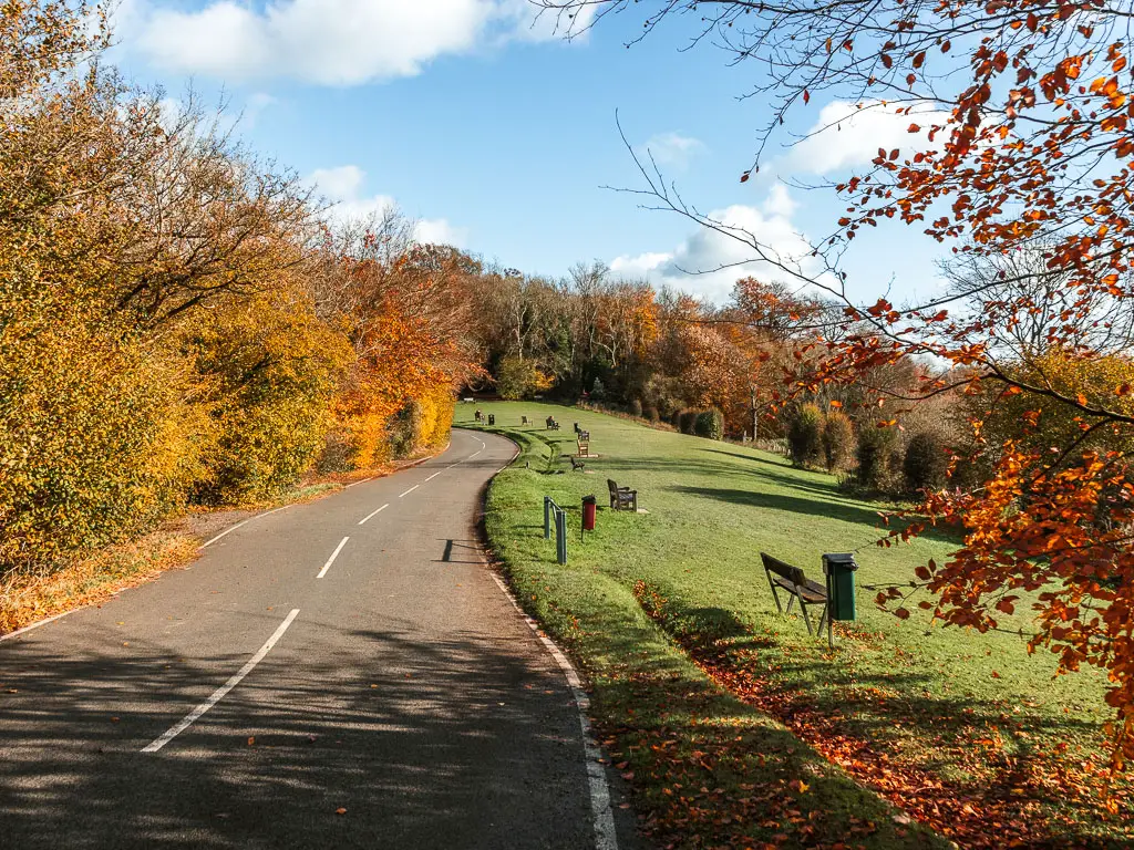 A road curving ahead, with a large green to the right, at the halfway point in the walk between Merstham and Oxted. The green has neatly cut grass with a row of wooden benches.