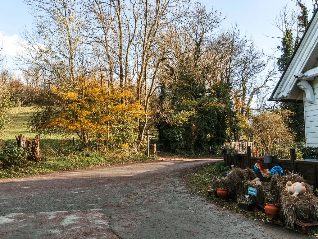 A road corner, with part of a white coloured house visible off frame to the right. There are trees lining thew road ahead. 