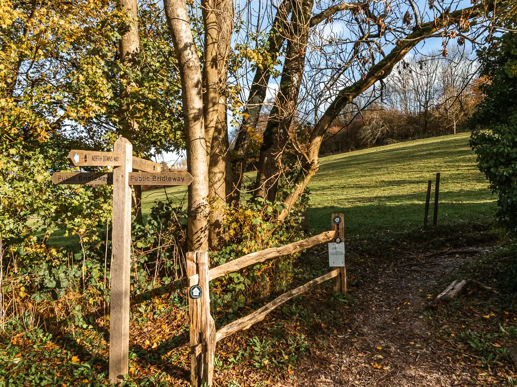 A wooden trail signpost pointing along the dirt trail leading up the hill. There is a wooden fence next to the sight, with trees behind it, and part of a green grass hill visible past the trees. 