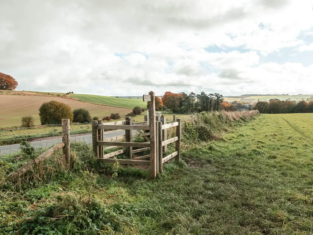 A wooden gate on the edge of the green grass field, leading onto the road on the left. 