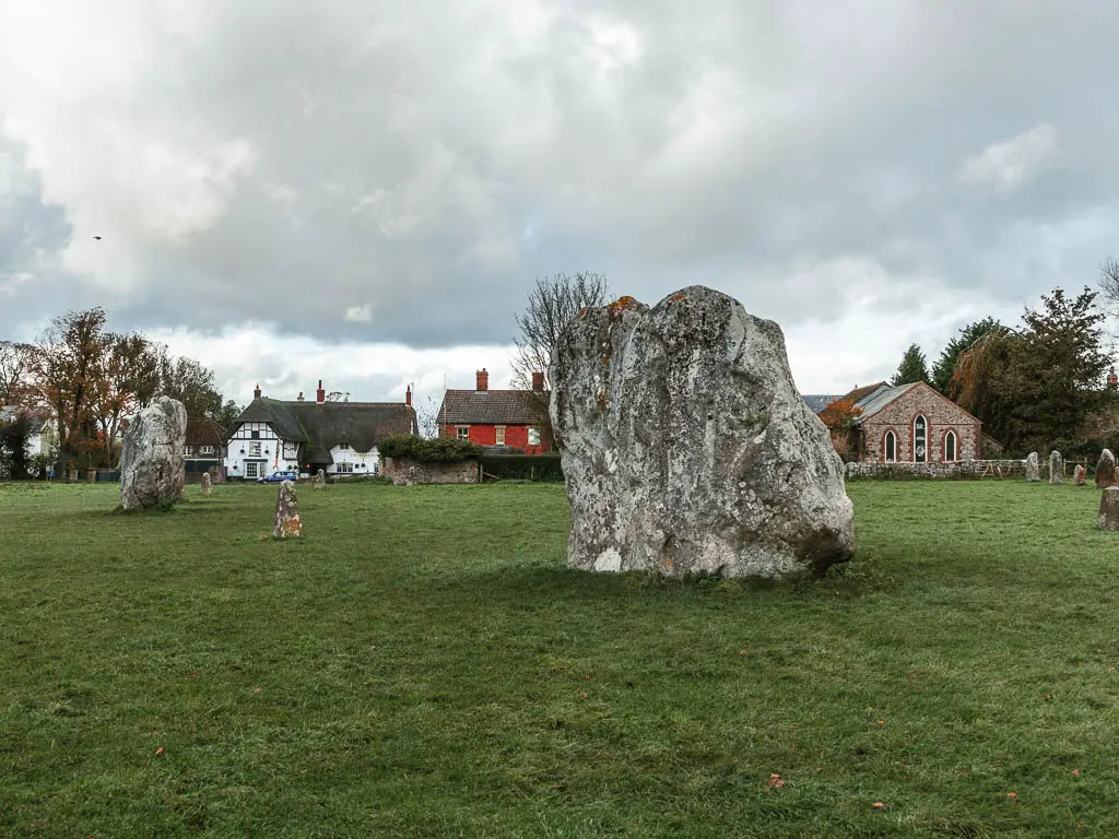 Looking across the green grass field to two large stones in Avebury, at the Strat of the walk towards Ogbourne St George. There are thatched roofed cottages with red and white walls on the other side of the field.