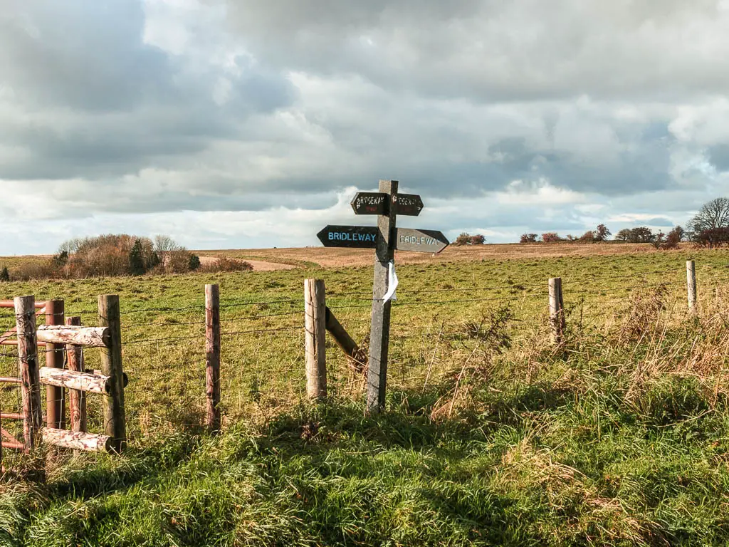 A wooden trail sign post pointing the way along the Ridgeway between Avebury and Ogbourne St George. The sign is in front of a wire fence with a green grass field on the other side.