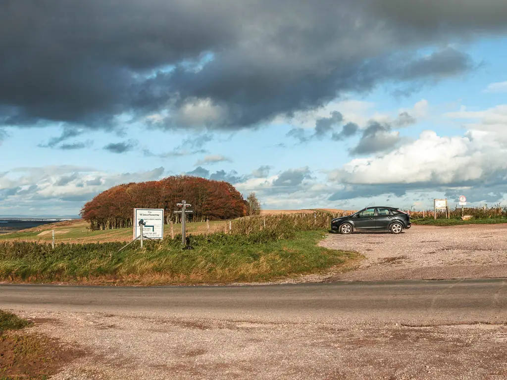 Looking across the road, to a car parking area on the other side, and one black cart parked. There is a group of red leafed trees ahead to the left. 