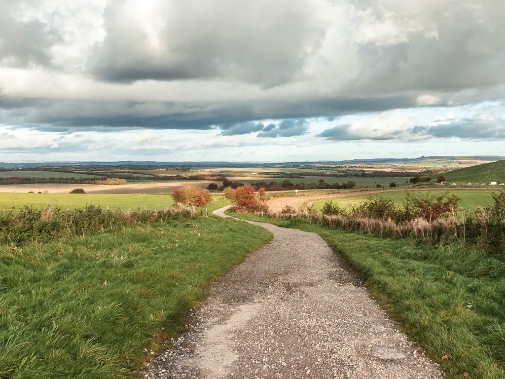 A gravel path winding ahead, surrounded by an expanse of grass and crop fields into the distance. 