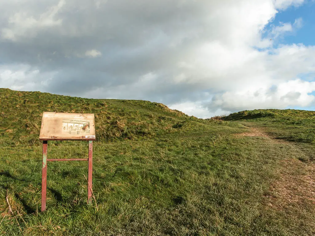 An information board on the left, in the grass, and a small hill behind it.