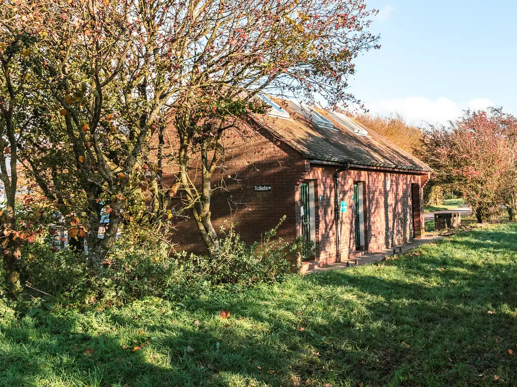 The brick walled toilet block, partially obscured by trees, standing next to the green.