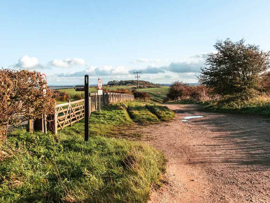 A dirt road on the right, and stip of grass and wooden gate and fence on the left. There is a trail signpost on the left side pointing through the gate.