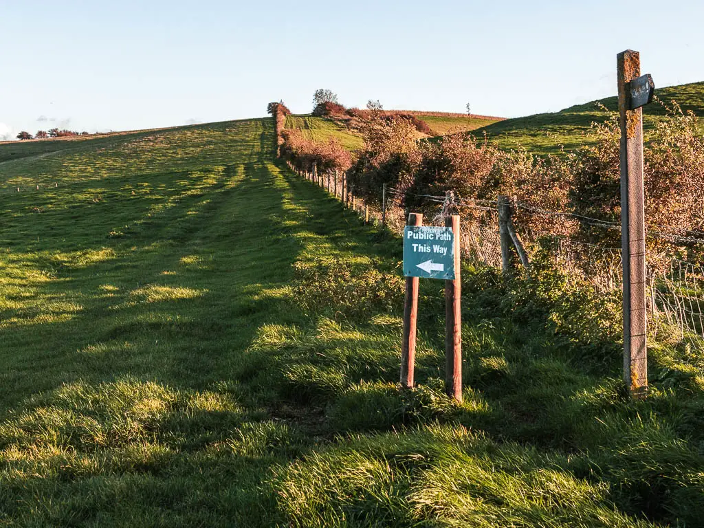 A grass field, with a fence and bushes on the right, and a public footpath sign pointing left.