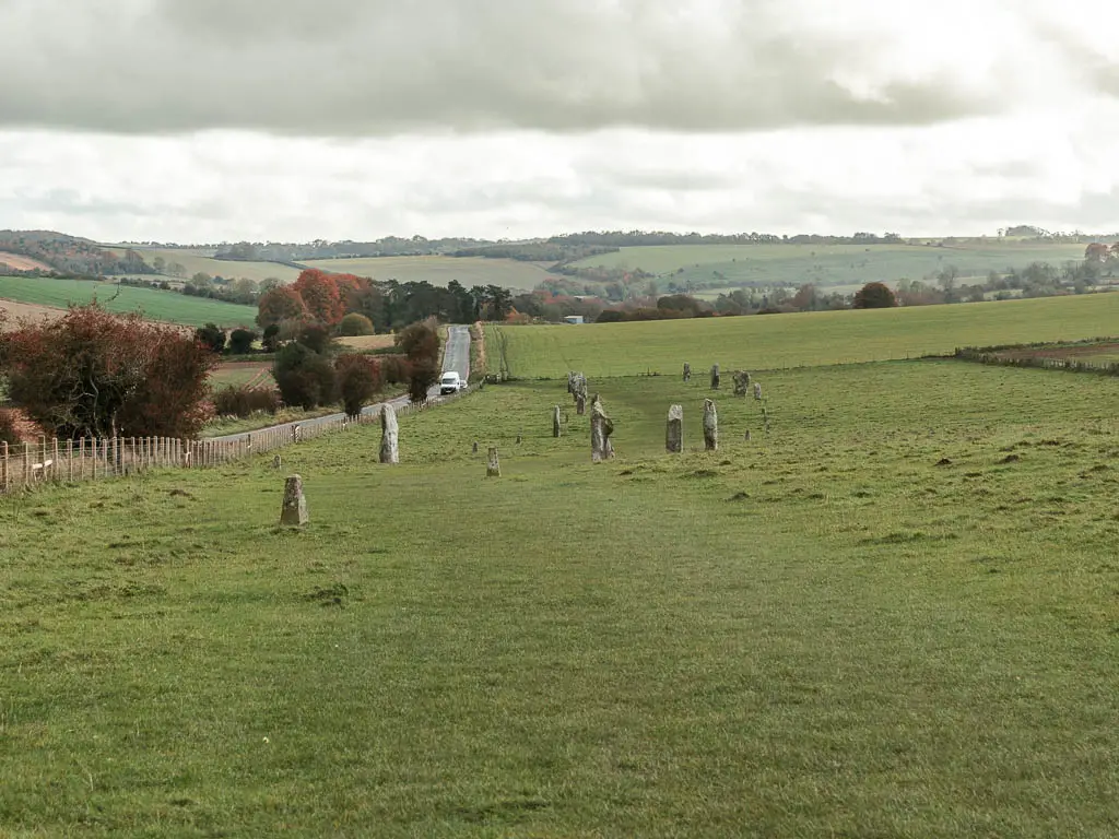 Looking across a large field with short grass, and a road running along the left side of it. There are standing stones ahead in the field. There are hills rising up in the distance.