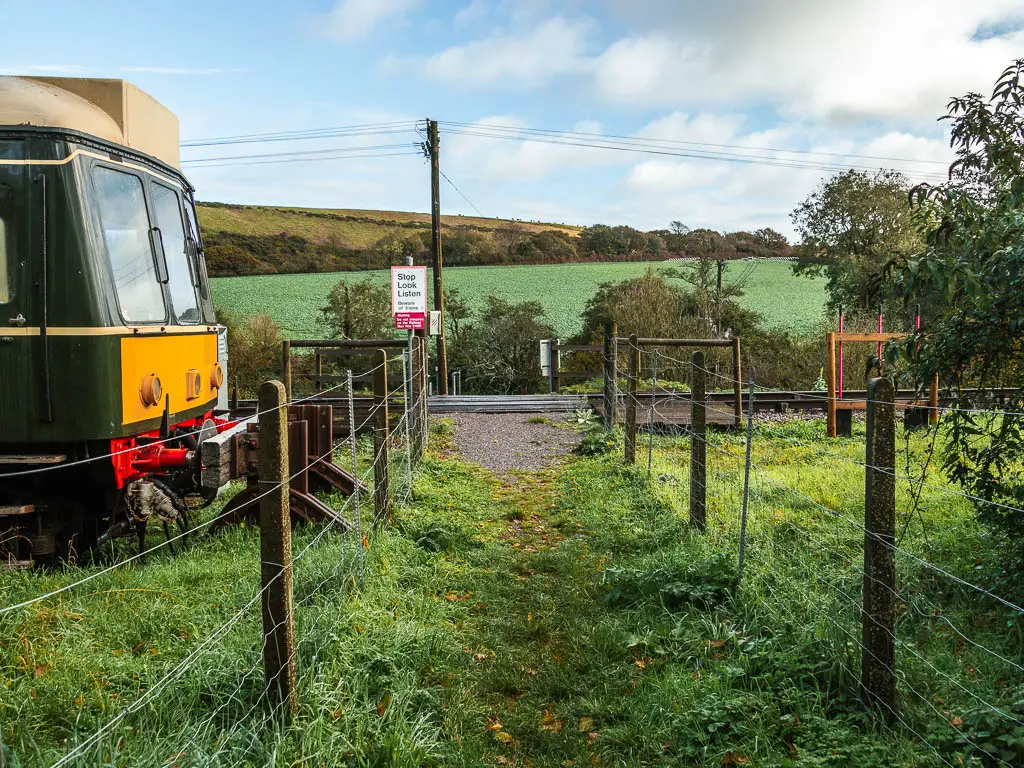 A grass path lined with metal fence, and a train carriage parked on the left, train tracks ahead, and a hill ahead in the distance. 