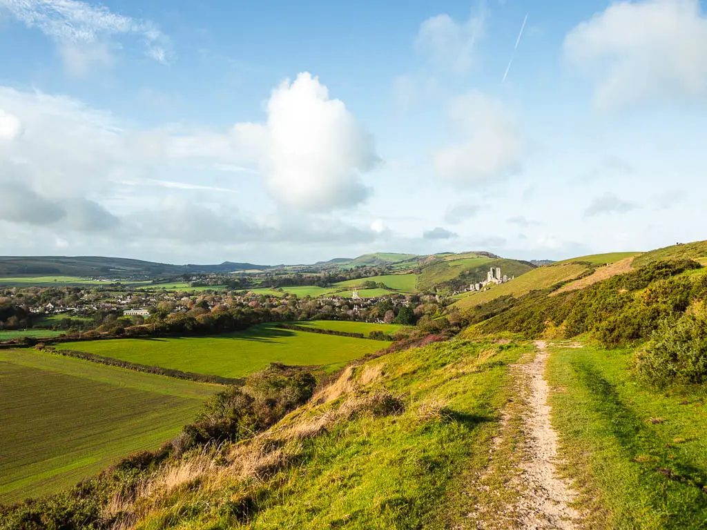 A dirt trail on the right, with a hill drop to the left, and a view to Corfe Castle in the distance on the walk towards Ballard Down and Swanage. there are large grass fields at the bottom of the hill.