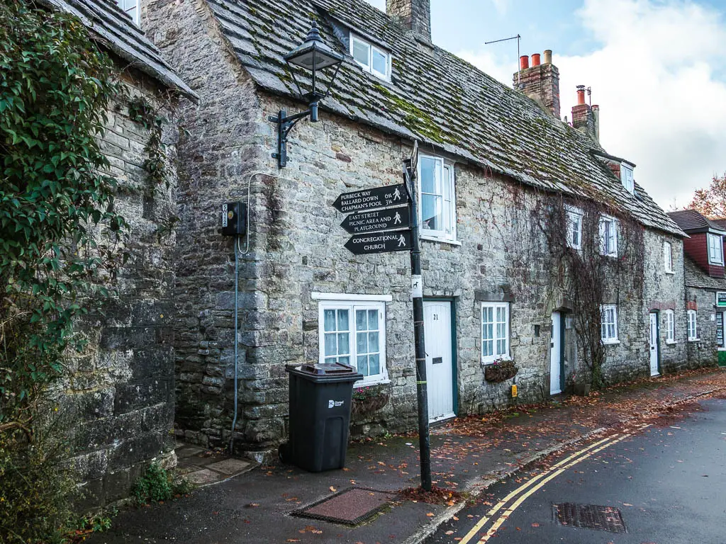 A black trail sign pointing left to walk towards Ballard Down along an alley between the stone houses in Corfe Village