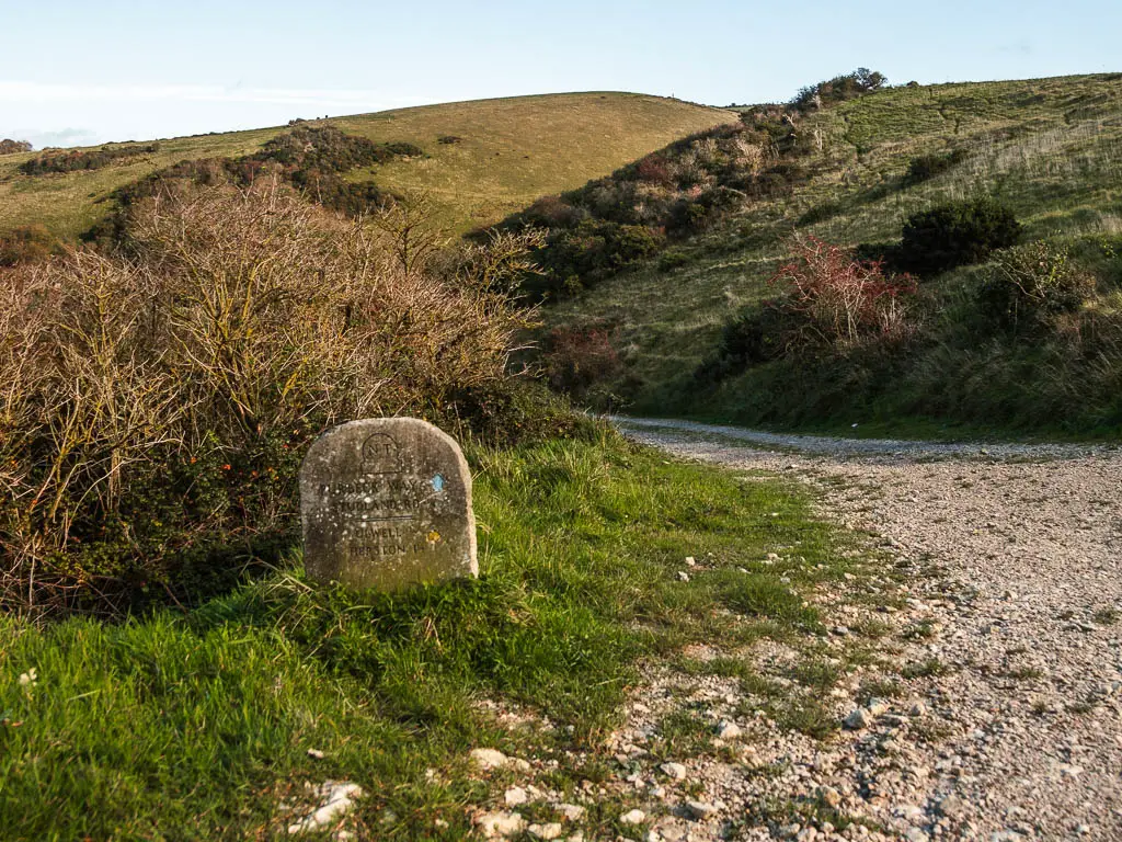 A stone trail marker on the left side of a gravel path, with a hill ahead on the other side.