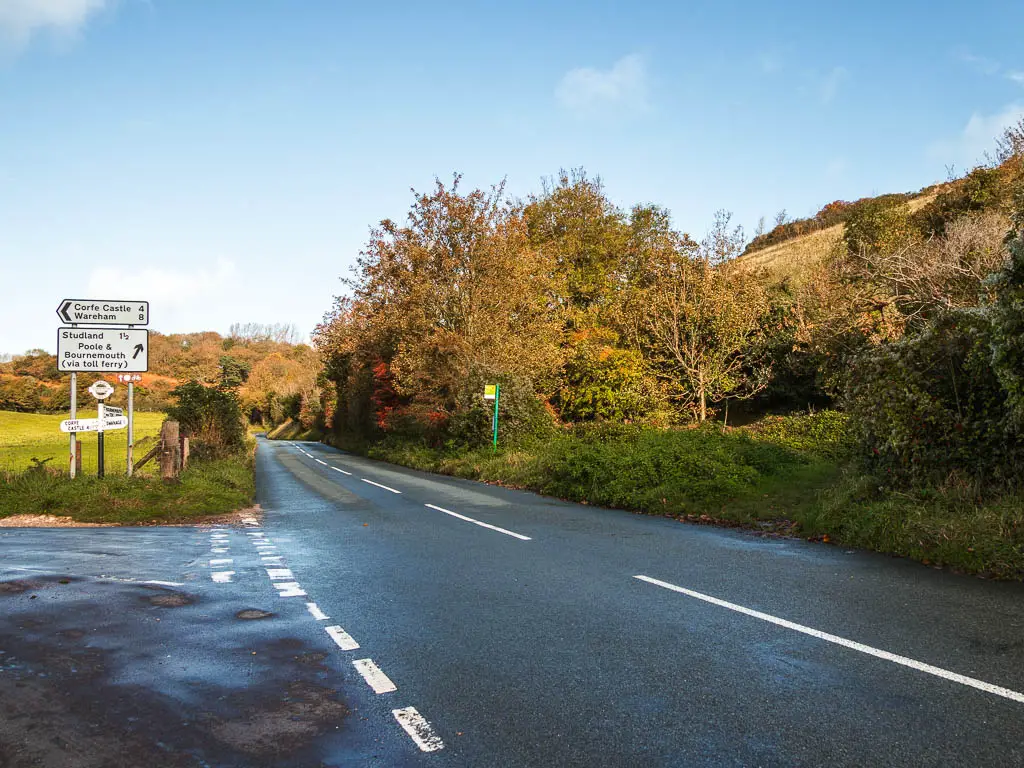 A road leading ahead, with bushes and trees on the right side, and a road sign on the left posing left to Corfe Castle.