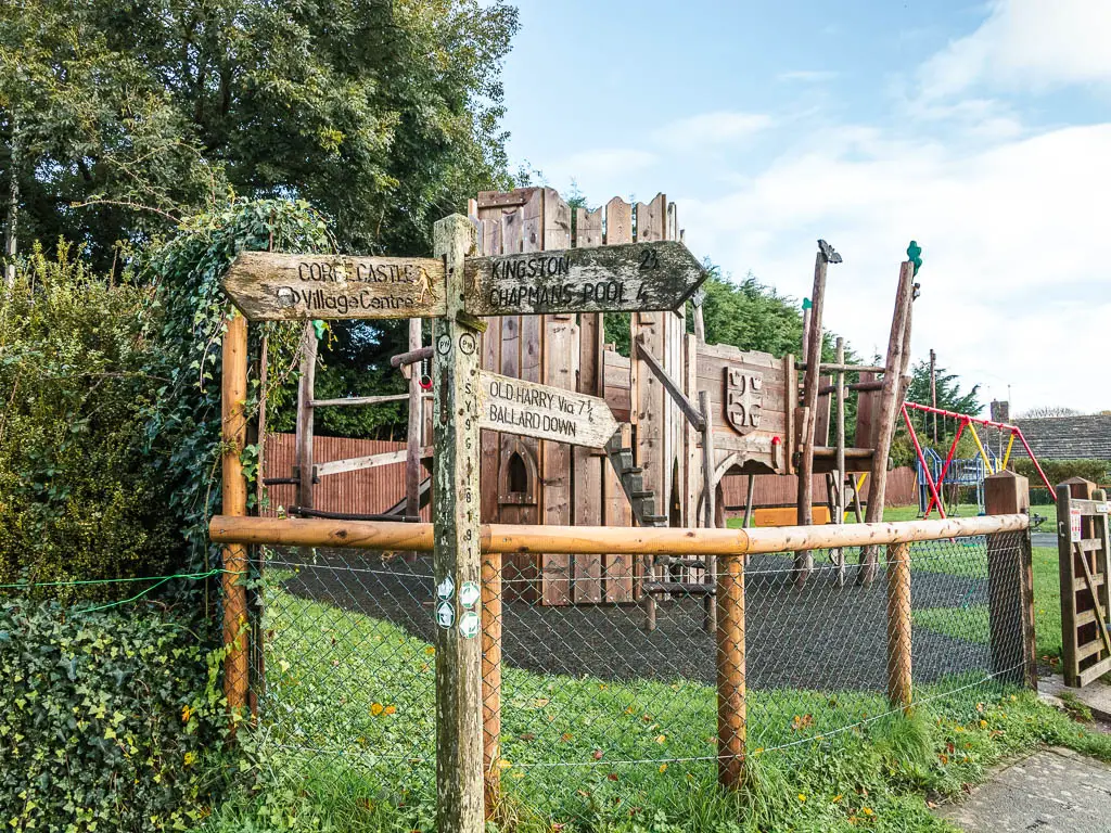 A wooden trail sign in front of a playground pointing left to walk to Corfe Castle and right to walk to Ballard Down.