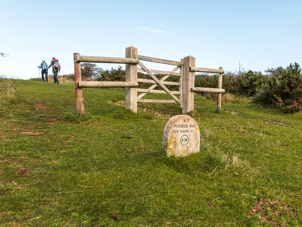 A stone trail sign saying 'Purbeck Way', infant of a wooden fence. there are two people walking ahead.