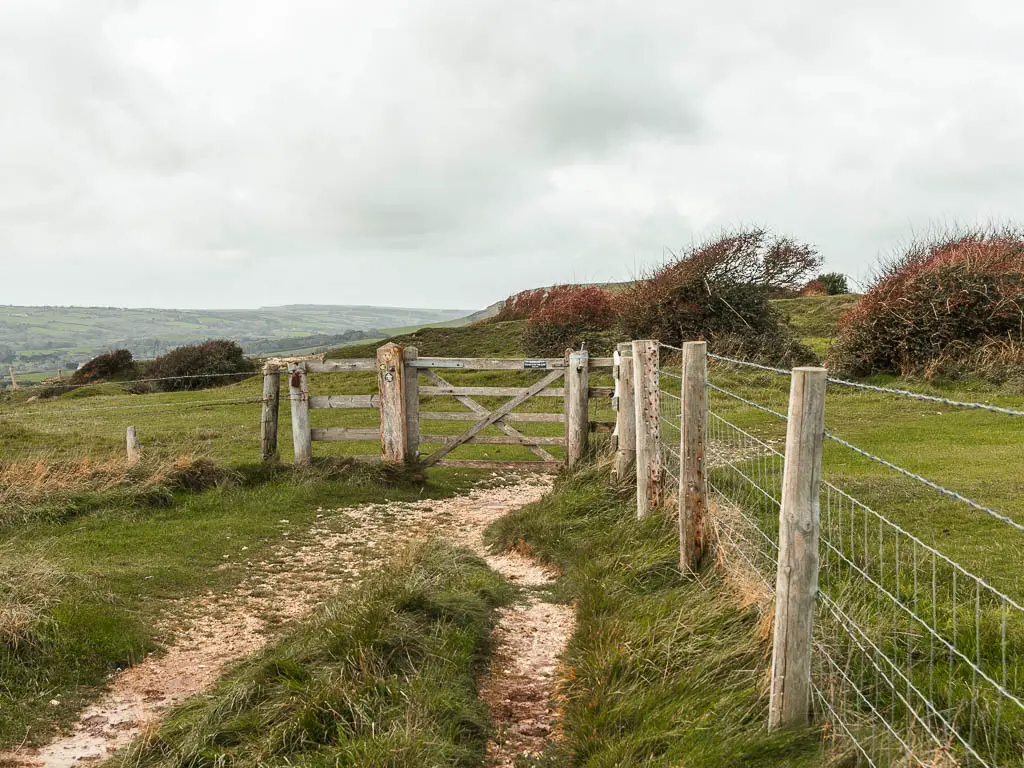 A dirt trail leading to a wooden gate. There is a wire and wooden fence on the right side.