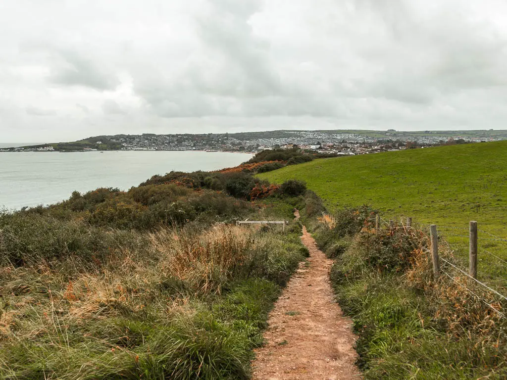 A dirt trial leading downhill with unkempt grass and bushes on the right and a green grass field on the left. There is a view towards Swnanage ahead, with the sea on the left.