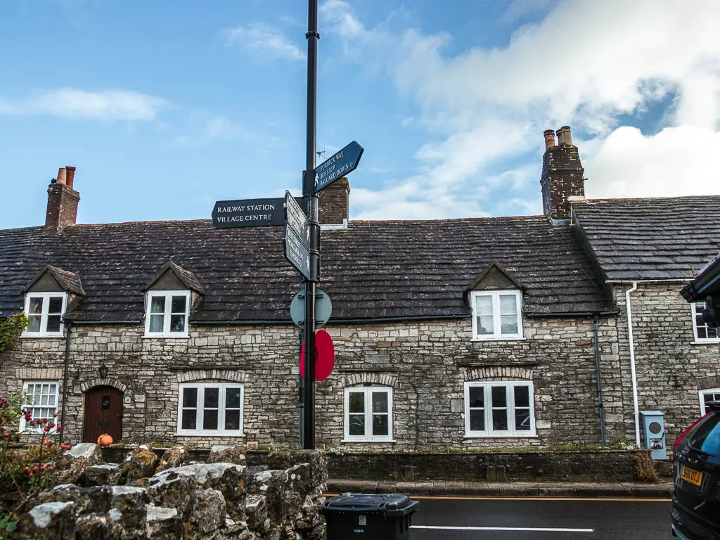 A tall metal pole with trail arrow signs pointing into a house to walk to Ballard Down, and back to walk to Corfe Castle. The pole is in front of a road, with stone houses on the other side.