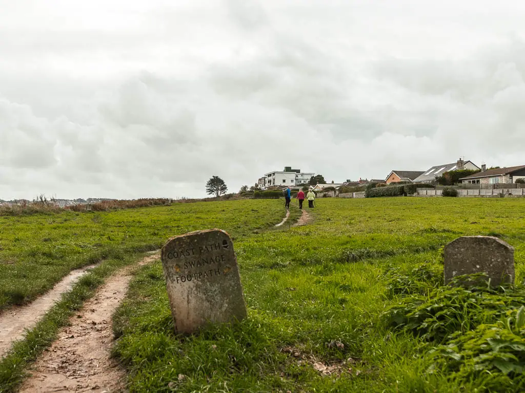 A stone trail sign next to the dirt trail leading through a large grass field. There are people walking along the trail ahead.