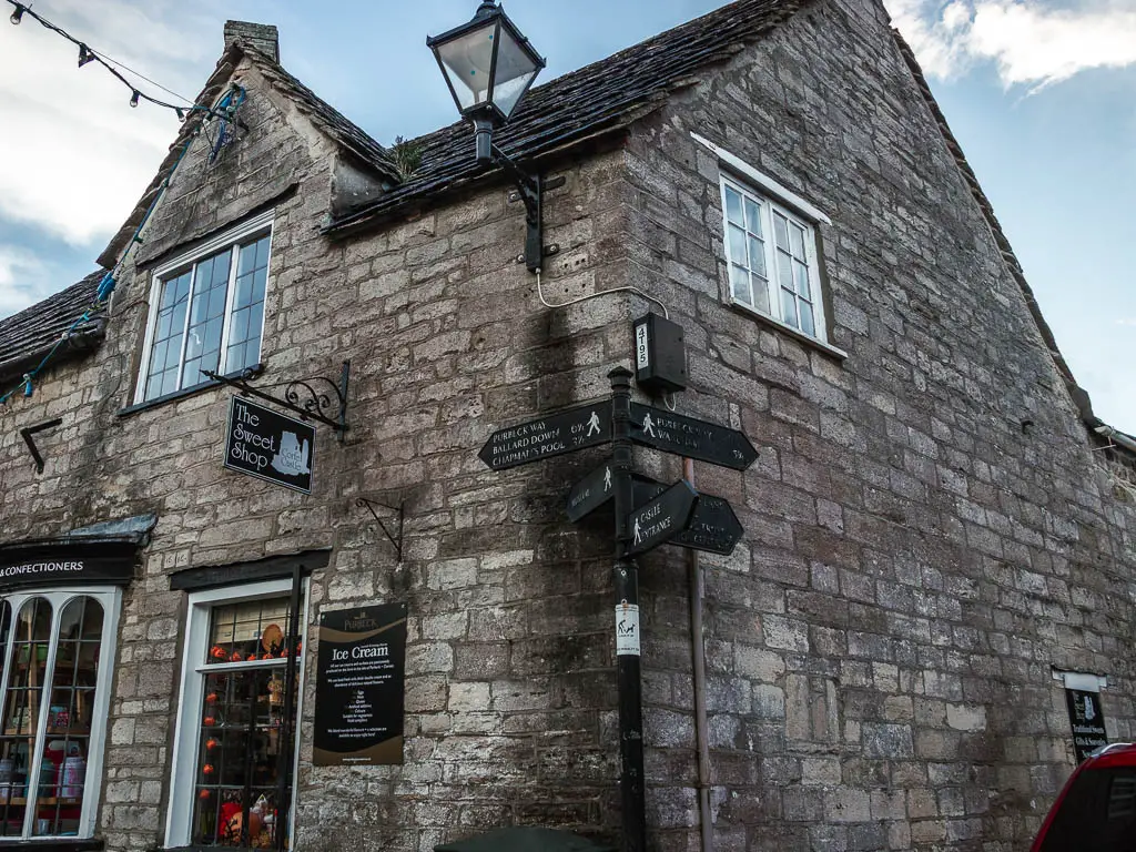 A black trail sign on the corner of a stone building in Corfe Village, pointing left to walk to Ballard Down and Swanage along the Purbeck Way.