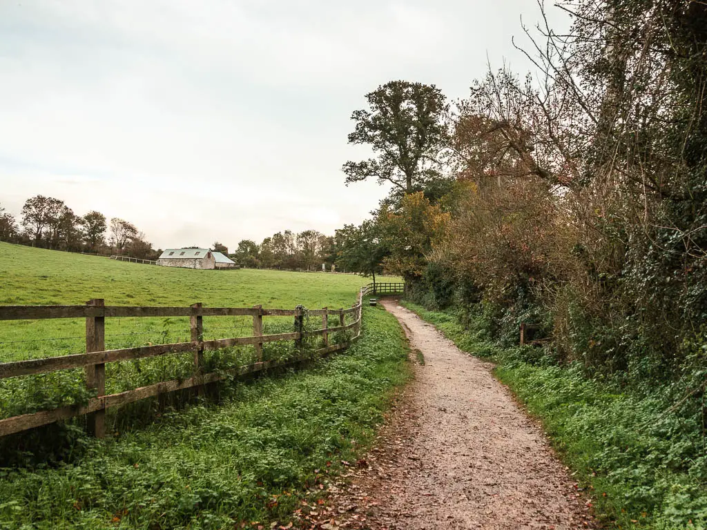 A walking path leading ahead, with bushes and trees on the right, and a wooden fence and green grass field on the left.